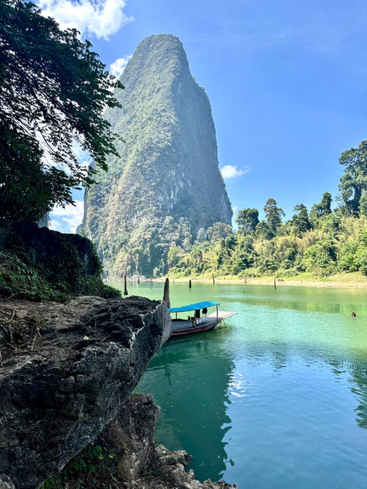 A blue-canopy longtail boat under a tall limestone tower on Cheow Lan Lake